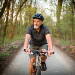 a senior man rides his bike through a paved path surrounded by trees