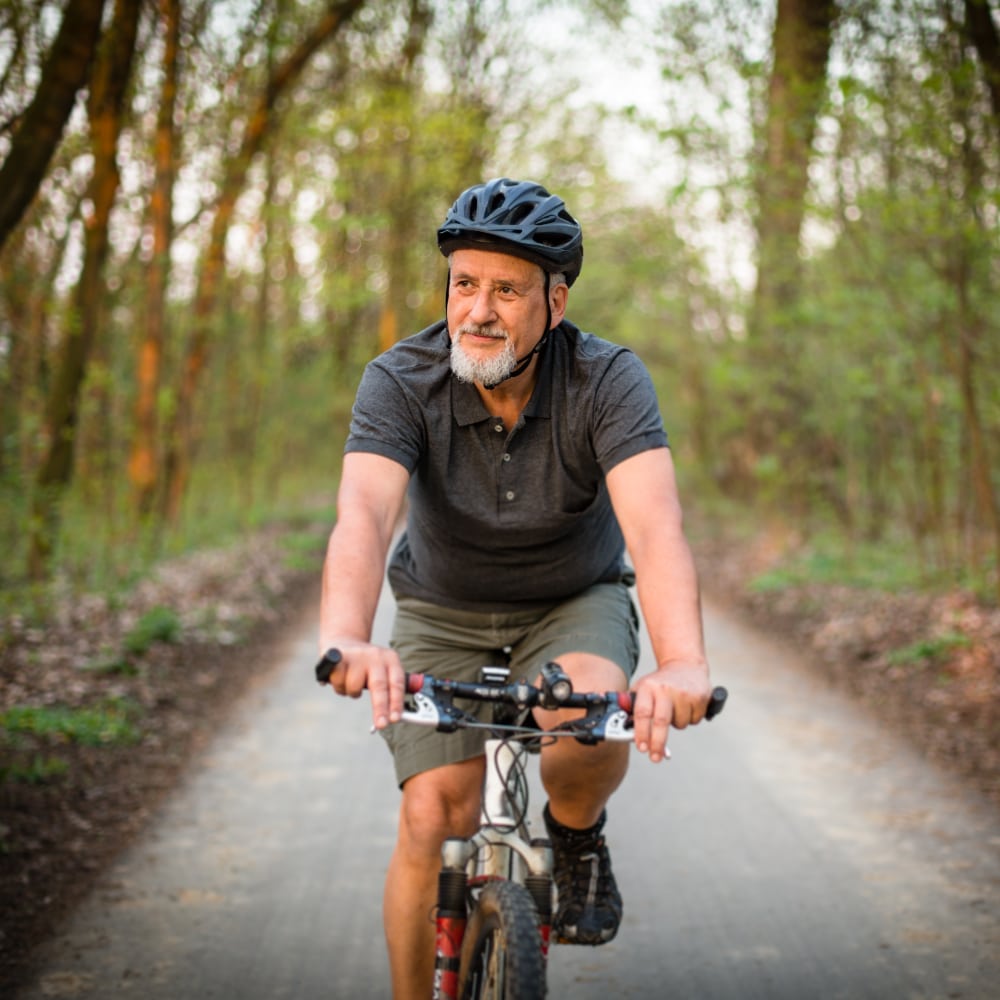 a senior man rides his bike through a paved path surrounded by trees