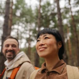Photo of a couple exploring the woods together. Shows faces, man to left looking left and smiling at woman who is looking straight ahead and smiling.